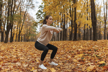 Full body of confident young female athlete in activewear squatting in autumn park while having fitness training in nature 