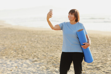 Smiling adult woman in training wear holding yoga mat make photo or have video call with smartphone over sea nature background. Middle aged blogger. Healthy lifestyle. Meditation, yoga and relaxation.