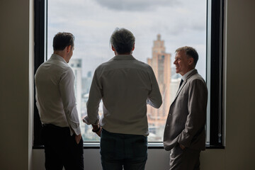 the backside of three businessmen deep in discussion together while standing in a high office building with windows overlooking the city, morning time.