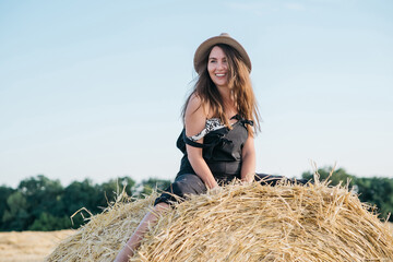 Naklejka na ściany i meble beautiful brunette woman in black overalls with a straw hat on a haystack in a field at sunset. High quality photo