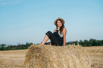 Naklejka na ściany i meble beautiful brunette woman in black overalls with a straw hat on a haystack in a field at sunset. High quality photo