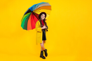 Happy teenager portrait. Teen girl under rainbow umbrella in autumn weather isolated on yellow background. Autumn kids clothes. Smiling girl in autumn coat.