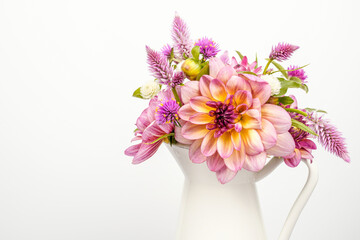 Bouquet of a variety of dahlias, celosia, and strawflower flowers. Arrangement is placed in a white vessel with a handle. White background. Space for text.
