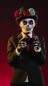 Vertical video: Model with santa muerte body art playing video games in studio, using controller to play on gaming console. Wearing spooky halloween costume of mexican day of the dead tradition.