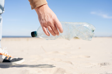 Volunteer man's hand picking up plastic bottle, collecting waste on sea beach.