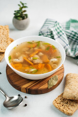 
chicken soup broth with vegetables in a white bowl, next to a spoon, bread, towel, spices
