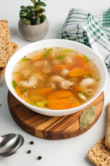 
chicken soup broth with vegetables in a white bowl, next to a spoon, bread, towel, spices