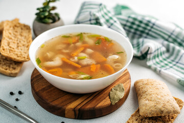 
chicken soup broth with vegetables in a white bowl, next to a spoon, bread, towel, spices