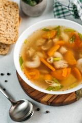 
chicken soup broth with vegetables in a white bowl, next to a spoon, bread, towel, spices