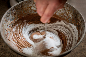 Close up of a hand mixing sugar and chocolate cake batter in a bowl.