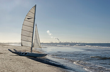Catamaran sailboat on an almost empty sandy beach near Monster, The Netherlands, with the cranes, chimneys and wind turbines of the Port of Rotterdam in the background