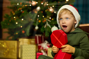 Child in a red hat, with big blue eyes and a mouth open in surprise tears off a beautifully packaged gift under a decorated Christmas tree.