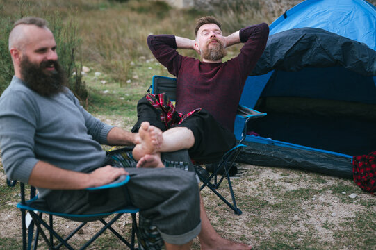 Hipster long beard gay couple camping on open grass field. Attractive male homosexuals relaxing on chairs in front of tent in late autumn. Man holding partner's  barefoot legs in his lap.