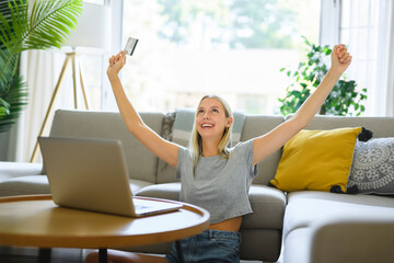 Happy teenager girl online with a laptop in the living room at home with credit card