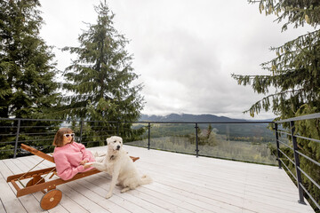 Woman in pink sweater rests with her dog on a spacious white terrace in mountains. Recreation in...