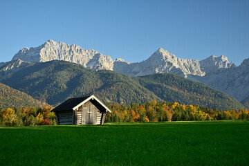 Fototapeta na wymiar Ein Heuschober steht auf einer grünen Wiese vor einem herbstlich bunten Wald und dem Karwendel Gebirge bei blauem Himmel.