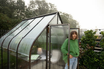 Portrait of young woman stands near greenhouse for growing plants at backyard. Gardening, leisure time in garden concept