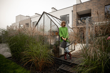 Portrait of young woman stands with watering can near glasshouse for growing plants and pampas...
