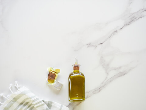 Small Glass Bottle With Olive Oil, Unwrapped Stock Cube And Tea Towel On White Marble Kitchen Counter, Top View With Copy Space.