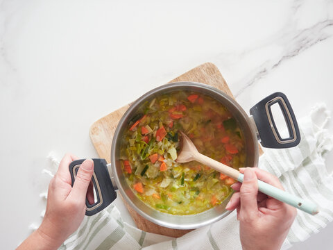 Female Hands Stirring Pot Of Vegetable Soup With Wooden Spoon. View From Above Marble Kitchen Countertop, Wooden Board And Dishcloth