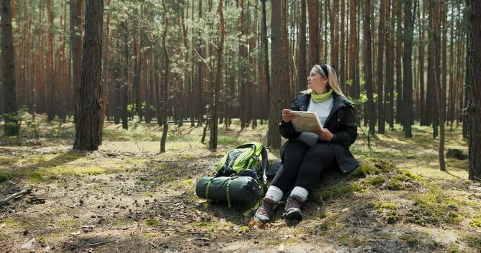 Photo from distance happy blonde woman with headband sitting on grass in middle of forest with travel backpack mat and special walking sticks looking for way to destination holding map in hands.