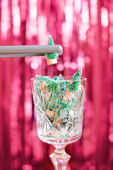 A vintage glass goblet filled with miniature Christmas trees against a backdrop of red glittery tinsel. The tongs add another tree to the glass. Humorous New Year or Christmas concept.