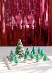 A miniature Christmas tree surrounded by toy trees is in a white frame against a background of red shiny tinsel. Abstract minimalistic christmas composition with copy space.