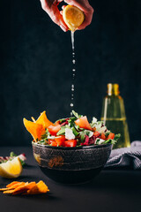 vegetable salad in a bowl on a dark background with a female hand that squeezes a lemon onto a dish
