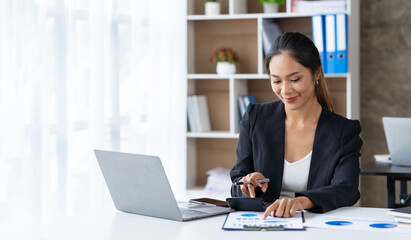 Beautiful Asian businesswoman analyzing charts using company earnings calculator. and laptop at the...
