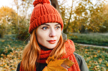 Portrait of mysterious woman with autumn yellow leaf in hand in orange knitted hat and scarf. Autumn inspiration.