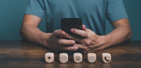 Contact Us concept, Man hand holding Mobile Phone for customer communication service with contact icon, E-mail, Phone number, Location, Social media group on the wooden box.
