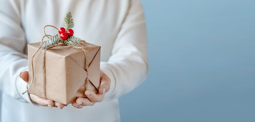 A woman a holds a craft paper box in her hands with a Christmas decor made from a fir branch and red berries. Close-up of female hands holding a Christmas or New Year's gift.