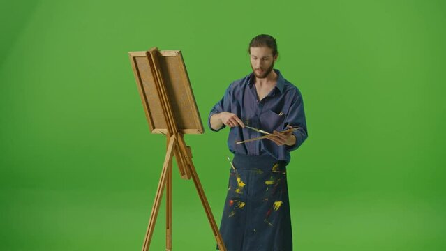 Young Talented Artist in Denim Shirt and Apron Working on Creation of New Painting.Motivated Bearded Man Draws with Brush on Canvas on Easel on Green Screen,Chroma Key.The Creativity And Art Concept