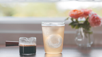 Iced coffee in a clear glass in the morning with natural light by the window. Green nature background and sweet pink roses, Healthy drink concept, Kombucha americano, Fermented tea with probiotics.