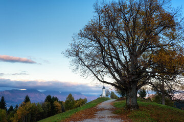 View of the Jamnik church St Primus and Felician at sunset, Alps mountains, Slovenia. Beautiful landscape with footpath, trees and blue sky with clouds, outdoor travel background