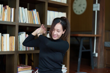 A portrait of a young Asian woman using a computer, wearing headphones and using a notebook to study online shows boredom and pain from video conferencing on a wooden desk in library