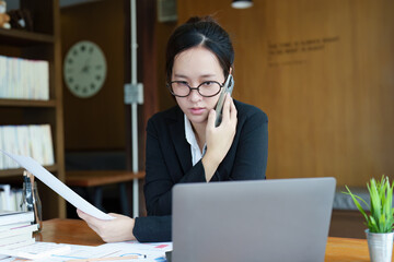 Portrait of a young Asian woman showing a serious face as she uses her phone, financial documents and computer laptop on her desk in the early morning hours