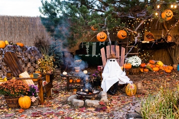 Little white halloween ghost in hat and glasses sits on wooden chair  by the fire. Halloween...