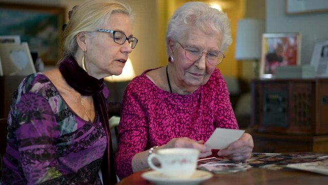 Slow motion of happy elderly woman and mature daughter having fun looking at old photo memories at the dining room table.