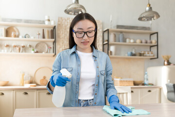 Maid services. Young beautiful Asian woman works as a cleaner. Cleans the house. Wipes the table with a cloth in rubber gloves, sprays the surface with a spray.