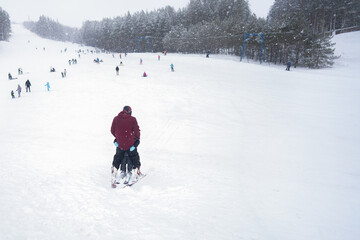 Ski resort in a snowfall. A cluster of skiers and snowboarders on a snow-covered slope. In the foreground, a man teaches a child to stand on downhill skis.