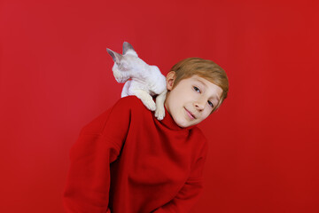 A cheerful boy in a red tracksuit with a small white kitten with blue eyes sitting on his shoulders climbs on the boy's shoulders