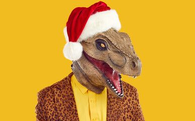 Portrait of man in funny unusual Christmas costume. Headshot of strange guy wearing bizarre dinosaur monster mask, red Santa Claus hat and funky leopard jacket isolated on yellow color background