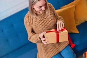 Photo of an adorable beautiful attractive cute cute girl showing you her wrapped present. A beautiful blonde woman in a sweater poses against the backdrop of Christmas lights with a gift in her hands.