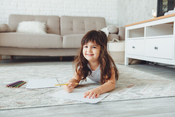 A cute little girl lies on the floor of the house and draws with pencils, children's leisure.