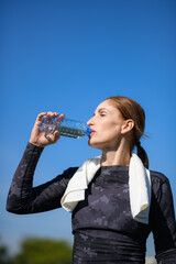 Women and sport. Girl in sportswear with a towel drinks water from a plastic bottle against the blue sky. Middle aged sportswoman dressed in sportsclothes