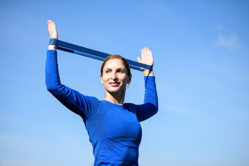 Women and sport. Smiling girl in sportswear - blue shirt and black leggings with a sports elastic band doing stretching against the sky. Close up middle aged sportswoman