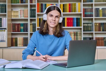 Portrait of teenage student guy sitting at desk in library, looking at camera