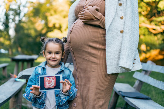 a little girl stands next to her pregnant mother as she holds and shows an ultrasound photo of her twin brother and sister.