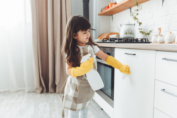 A cute little girl in an apron wipes the cupboard in the kitchen, washes with a sponge in a...